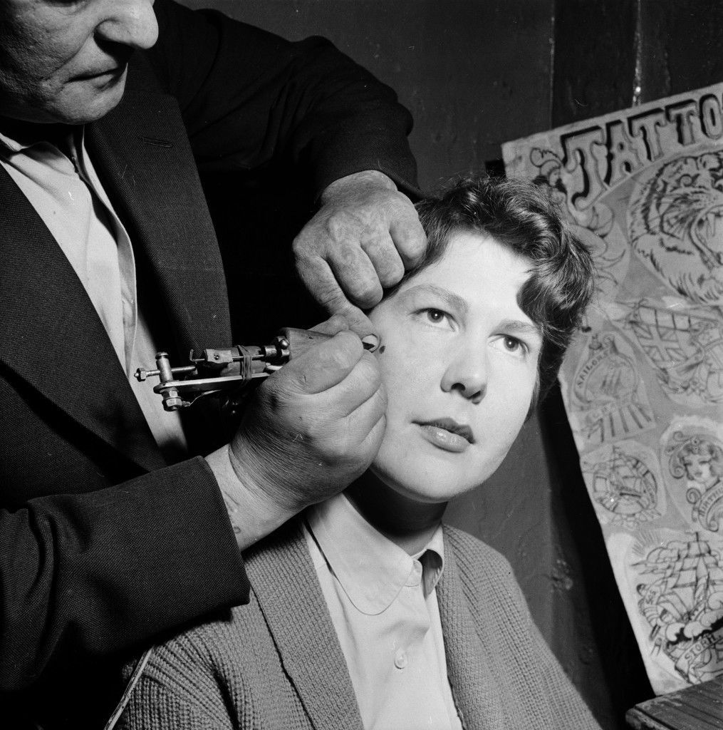 May 1956: A tattoo artist paints a permanent beauty spot onto the cheek of a female client at his workshop in Copenhagen. (Photo by John Firth)