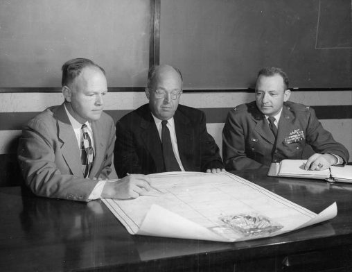 A group of men sitting at a table with a map, February 23, 1955. Shown above are the three officials of the Joint AEC-DOD Test Organization holding primary responsibility for conduct of the Spring 1955 nuclear tests at Nevada Test Site.